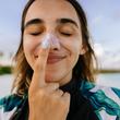 Photo of a young woman applying sunscreen while being outdoors, at the beach; making silly faces while reapplying SPF and protecting her skin from the sun.