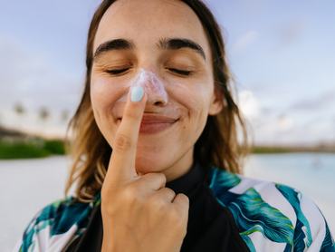 Photo of a young woman applying sunscreen while being outdoors, at the beach; making silly faces while reapplying SPF and protecting her skin from the sun.