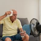Senior man sitting on sofa wiping sweat from forehead while holding glass of water near ventilator