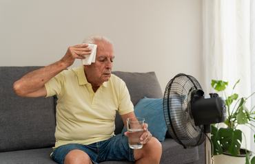 Senior man sitting on sofa wiping sweat from forehead while holding glass of water near ventilator