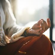 A close-up of a person meditating, wearing neutral-toned clothing, bathed in soft morning sunlight. The hand in a mudra gesture conveys calm and mindfulness.