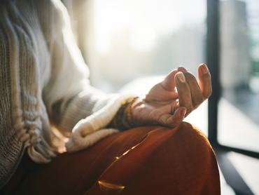 A close-up of a person meditating, wearing neutral-toned clothing, bathed in soft morning sunlight. The hand in a mudra gesture conveys calm and mindfulness.