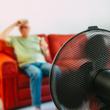 Egy hetvenes éveiben járó idős férfi ül a kanapén egy elektromos ventilátor előtt a nyári hőhullám alatt.