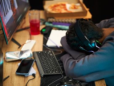 Young adult Black man wearing headphones sleeping at desk with head resting on arms near computer monitor, keyboard, smartphone, game controller, pizza box and drink visible on table